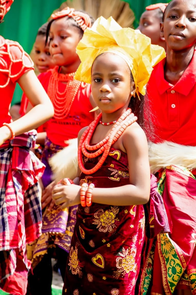 Young girl in traditional Nigerian attire with yellow headwrap and red beaded necklace, surrounded by other children in colorful traditional dress