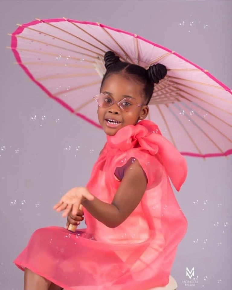 Young child wearing pink outfit and glasses holds a large pink umbrella against a gray background