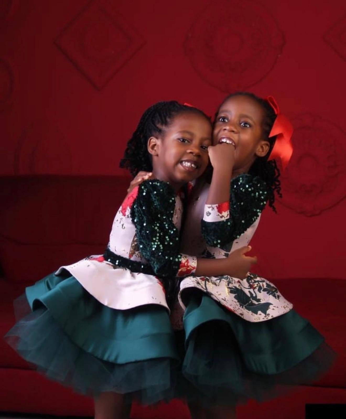 Two young girls in festive holiday dresses with sequin details and teal tulle skirts, smiling joyfully against a red background