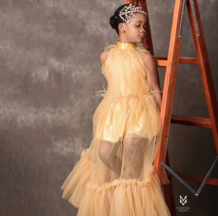Young girl in a peach tulle dress with tiered ruffles and a jeweled headpiece, posing next to an orange ladder against a gray backdrop