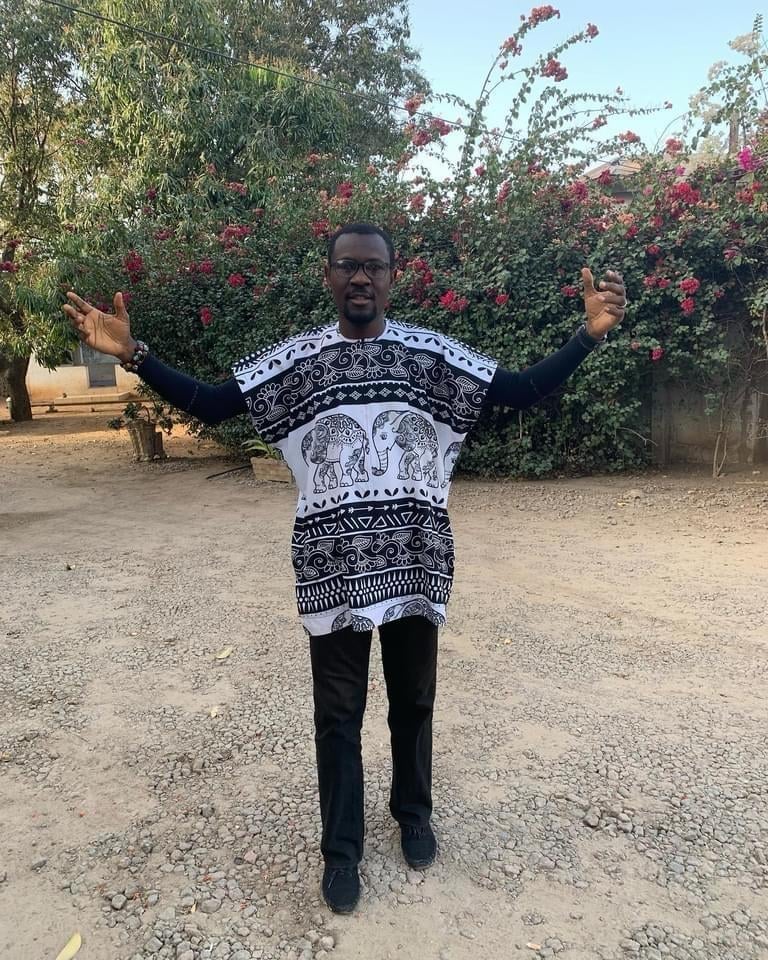 Man wearing a black and white elephant-patterned sweater with arms raised, standing on a dirt path with blooming flowers in the background.
